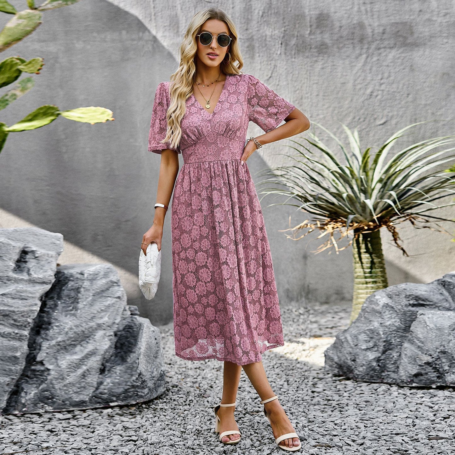 Donna con abito rosa in pizzo, occhiali da sole e sandali bianchi, posa tra piante e rocce. Moda estiva elegante, look casual chic.