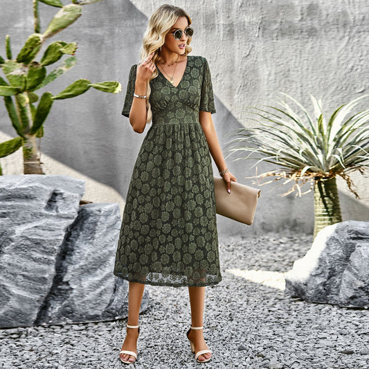 Donna elegante con abito verde floreale, occhiali da sole e borsa beige, posa in giardino moderno con piante succulente e pietre decorative.