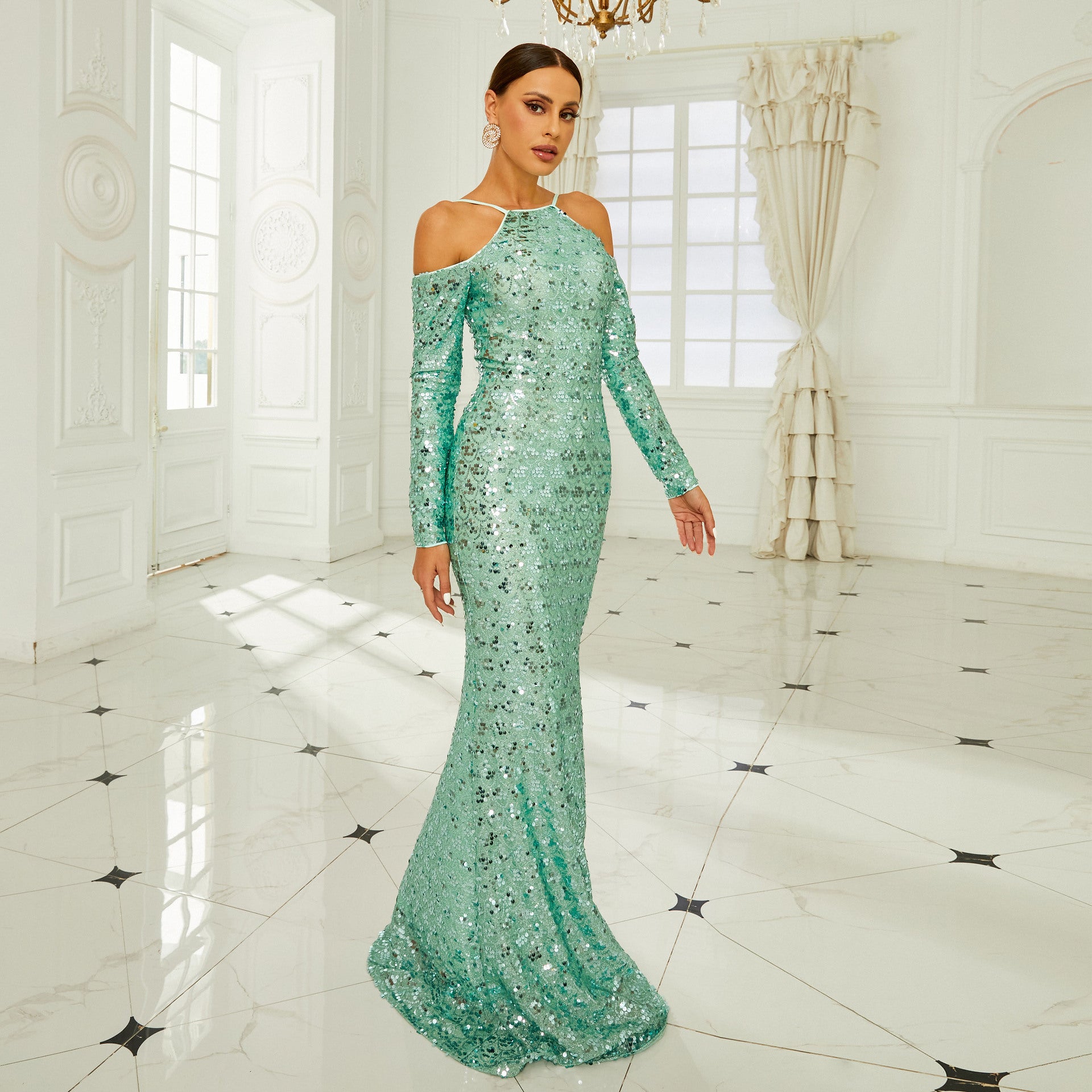 Donna in abito da sera verde brillante con paillettes, posa in elegante salone bianco con pavimento in marmo e grandi finestre. Moda, eleganza, lusso.