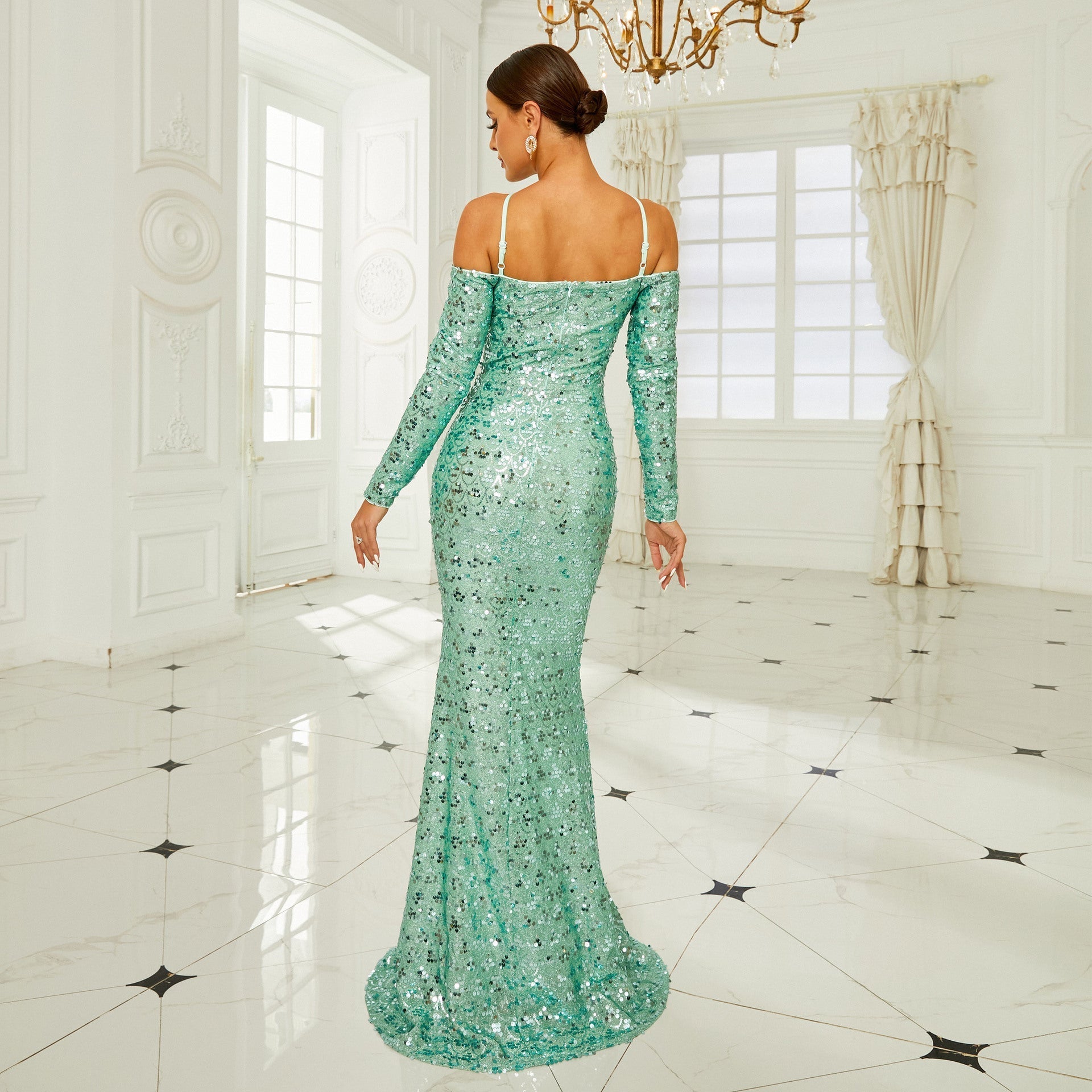 Donna in abito lungo verde smeraldo con paillettes, in una stanza elegante con pavimento in marmo e lampadario. Moda, eleganza, abito da sera.