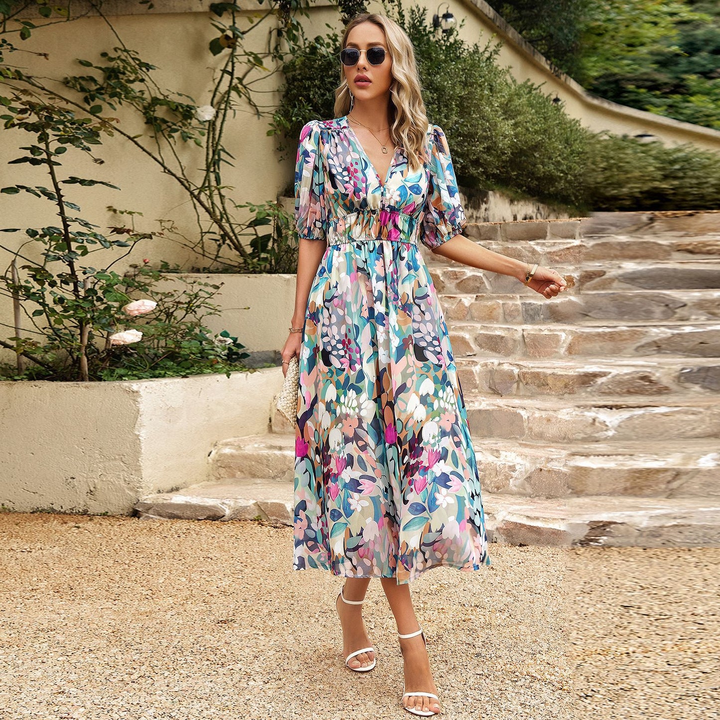 Donna con abito floreale lungo, occhiali da sole e sandali bianchi su gradini di pietra, circondata da piante verdi. Moda estiva elegante.