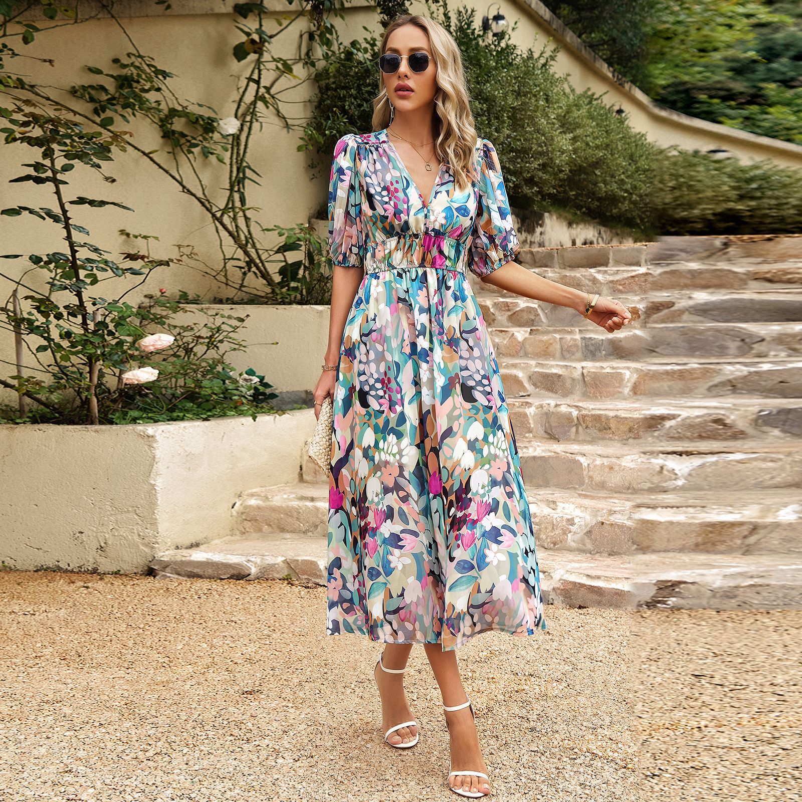 Donna con abito floreale lungo, occhiali da sole e sandali bianchi su gradini di pietra, circondata da piante verdi. Moda estiva elegante.