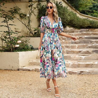 Donna con abito floreale lungo, occhiali da sole e sandali bianchi su gradini di pietra, circondata da piante verdi. Moda estiva elegante.
