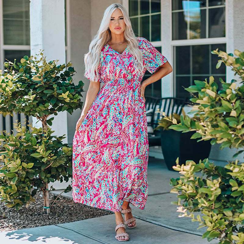 Donna con abito lungo floreale colorato, posa all'aperto vicino a piante verdi. Moda estiva, abbigliamento femminile elegante, stile casual chic.