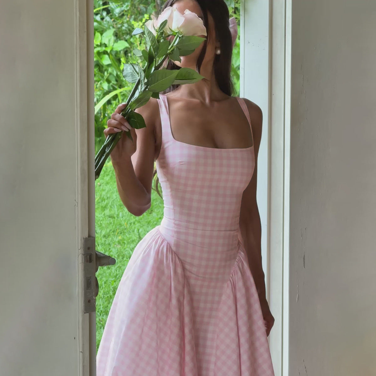 Donna in abito a quadretti rosa chiaro, tiene un fiore bianco davanti al viso, in piedi su una soglia con sfondo di giardino verde.