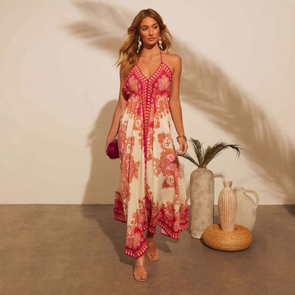 Donna in abito estivo lungo con stampa floreale rosa e bianca, sandali trasparenti, borsa fucsia, accanto a vasi decorativi su sfondo neutro.