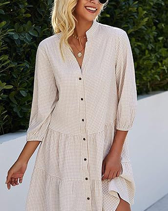 Abito estivo a quadri beige con bottoni, maniche a tre quarti, indossato da donna sorridente con occhiali da sole, sfondo di piante verdi.