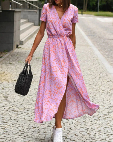 Donna con abito lungo floreale rosa e viola, cammina su strada lastricata, borsa intrecciata nera, scarpe da ginnastica bianche. Moda estiva casual.
