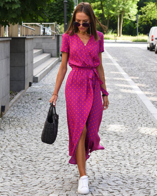 Donna con abito lungo fucsia a pois, borsa nera intrecciata, occhiali da sole, cammina su strada acciottolata. Moda estiva, stile casual chic.