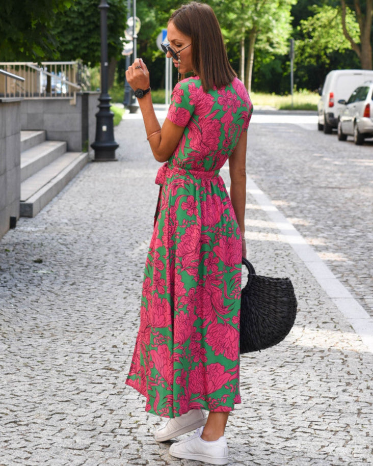 Donna con abito floreale rosa e verde, borsa nera intrecciata, cammina su strada acciottolata. Moda estiva, look casual, scarpe da ginnastica bianche.