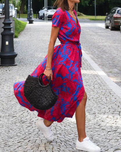 Donna con abito floreale rosso e blu, borsa nera intrecciata, cammina su strada acciottolata, indossa scarpe da ginnastica bianche.
