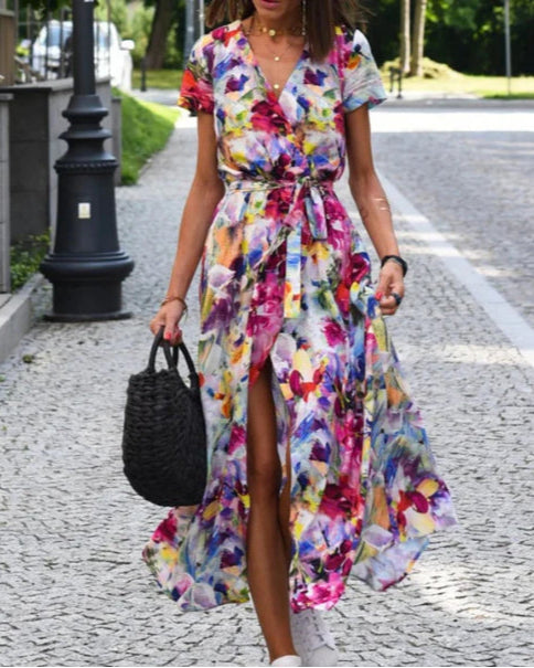 Donna con abito floreale colorato, lungo e con spacco, cammina su strada pavimentata. Borsa nera intrecciata, lampione e alberi sullo sfondo.