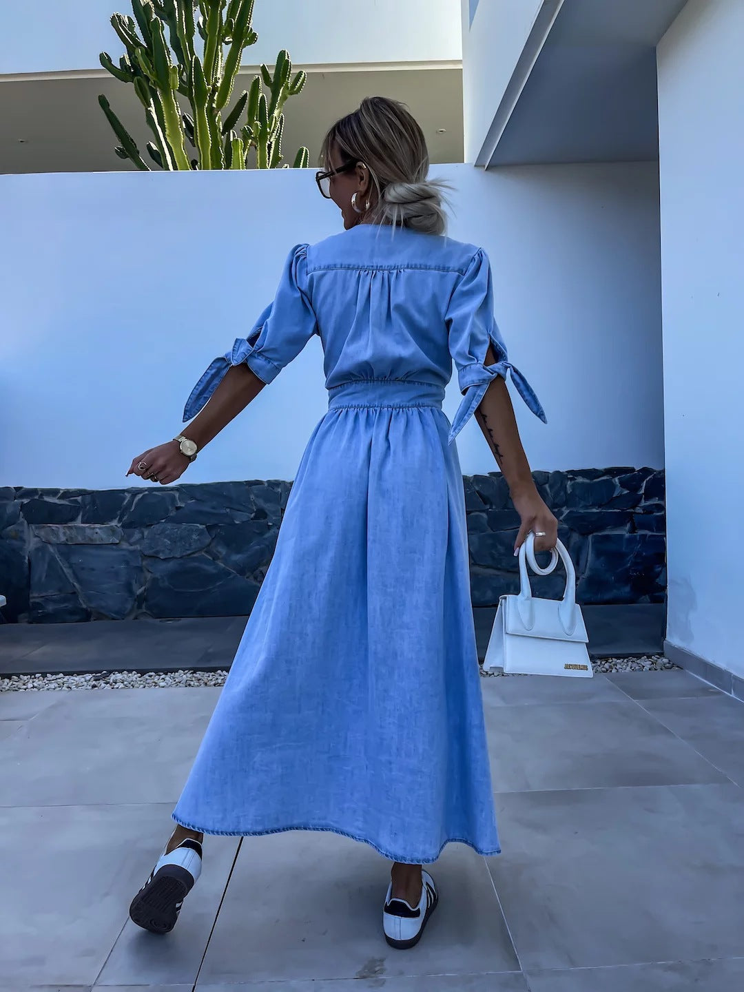 Donna di spalle con abito lungo in denim azzurro, maniche a fiocco, borsa bianca, scarpe casual, sfondo moderno con cactus. Moda estiva casual.