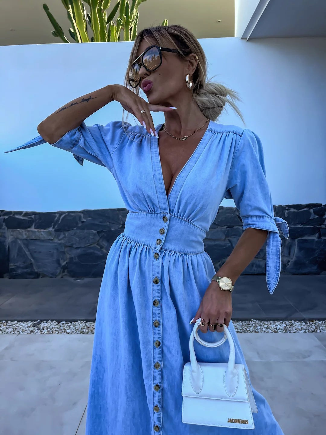 Donna con abito di jeans azzurro, occhiali da sole e borsa bianca Jacquemus. Sfondo con muro bianco e pianta di cactus. Moda estiva elegante.