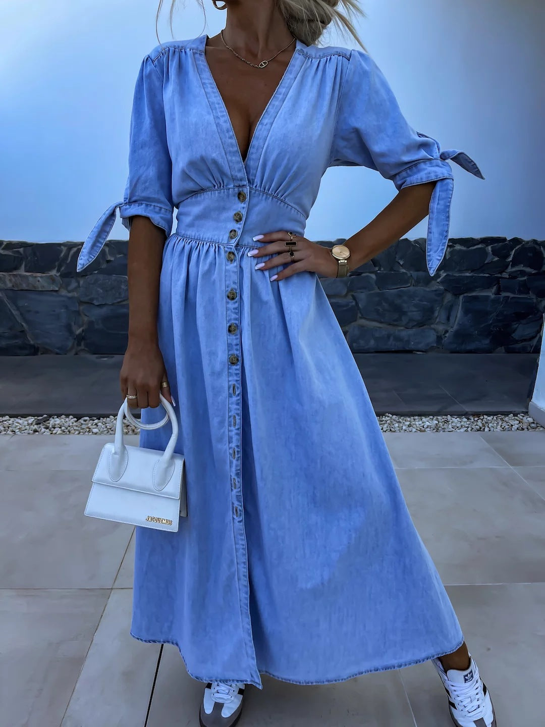 Donna con abito lungo in denim azzurro, bottoni frontali, maniche a tre quarti con fiocchi, borsa bianca piccola, orologio dorato, sneakers bianche.