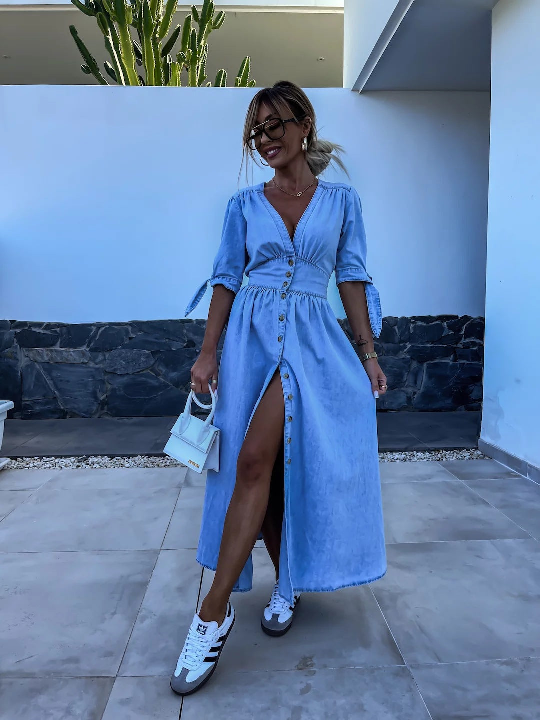 Donna in abito lungo di jeans con spacco, scarpe da ginnastica bianche e borsa piccola bianca, posa all'aperto davanti a un muro bianco e piante.
