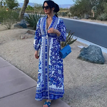 Donna con abito lungo blu e bianco a motivi floreali, borsa intrecciata blu, sandali blu, su marciapiede in ambiente desertico.