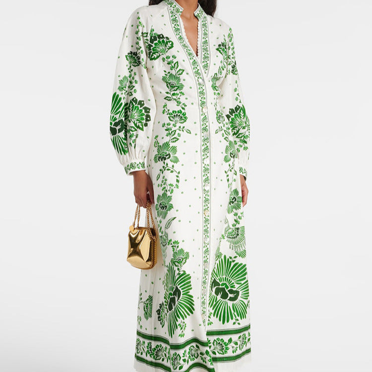 Abito lungo bianco con stampa floreale verde, maniche lunghe e colletto, indossato da una donna. Borsa dorata a mano. Moda elegante e sofisticata.