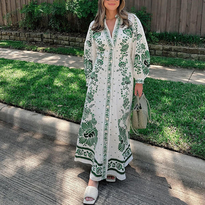 Donna con abito lungo bianco e verde a motivi floreali, borsa intrecciata verde, sandali bianchi, su marciapiede con erba e recinzione in legno.