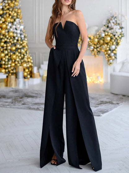 Donna in elegante tuta nera senza spalline, sfondo natalizio con alberi decorati e luci dorate, atmosfera festiva e sofisticata.