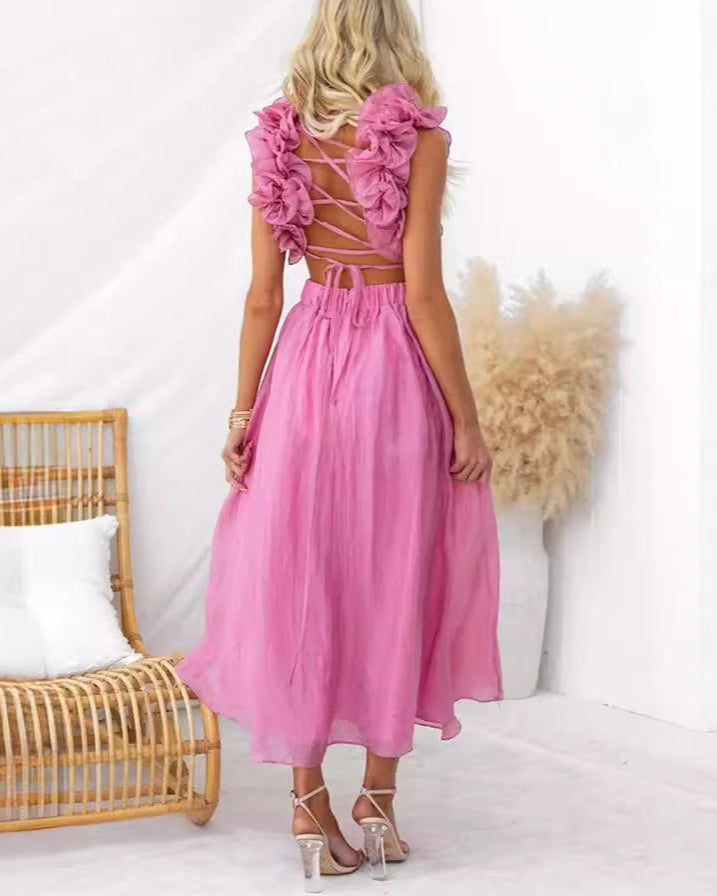Donna in abito rosa elegante con dettagli floreali sulla schiena, in piedi accanto a sedia di vimini e decorazione di pampas. Moda estiva femminile.