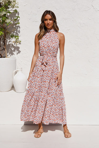 Donna in abito lungo floreale estivo, sfondo bianco, sandali marroni, vaso decorativo, moda femminile, abbigliamento casual elegante.