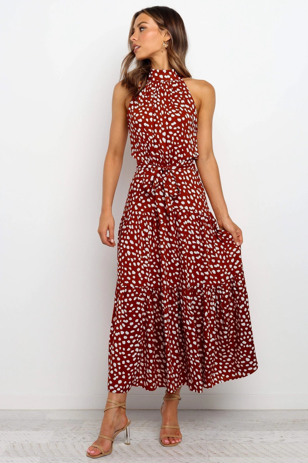 Donna con abito lungo rosso a pois bianchi, senza maniche, elegante e estivo. Indossa sandali dorati. Sfondo bianco minimalista.