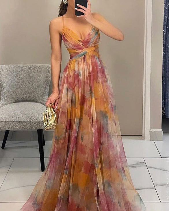 Abito lungo elegante multicolore con spalline sottili, motivo astratto, indossato da donna in piedi accanto a sedia grigia, borsa dorata in mano.