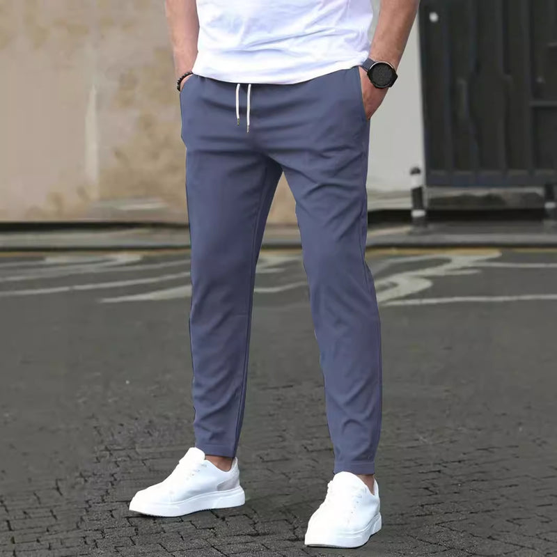 Pantaloni sportivi grigi da uomo, abbinati a scarpe da ginnastica bianche e maglietta bianca, su sfondo urbano. Moda casual maschile.