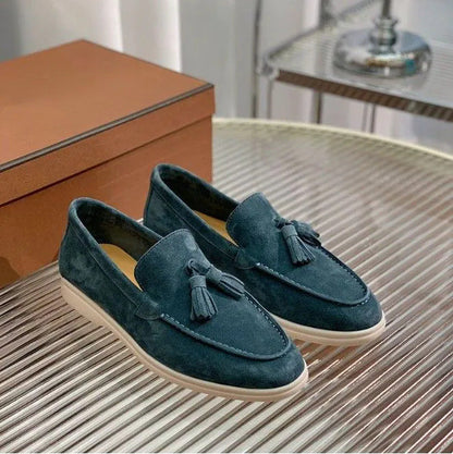 Mocassini in pelle scamosciata blu con nappine su tavolo di vetro, scatola marrone sullo sfondo. Scarpe eleganti uomo, moda calzature.
