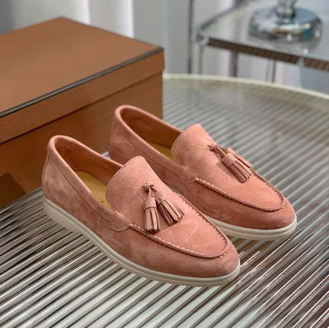 Mocassini rosa in pelle scamosciata con nappine su tavolo di vetro, scatola marrone sullo sfondo. Scarpe eleganti da donna, moda calzature.