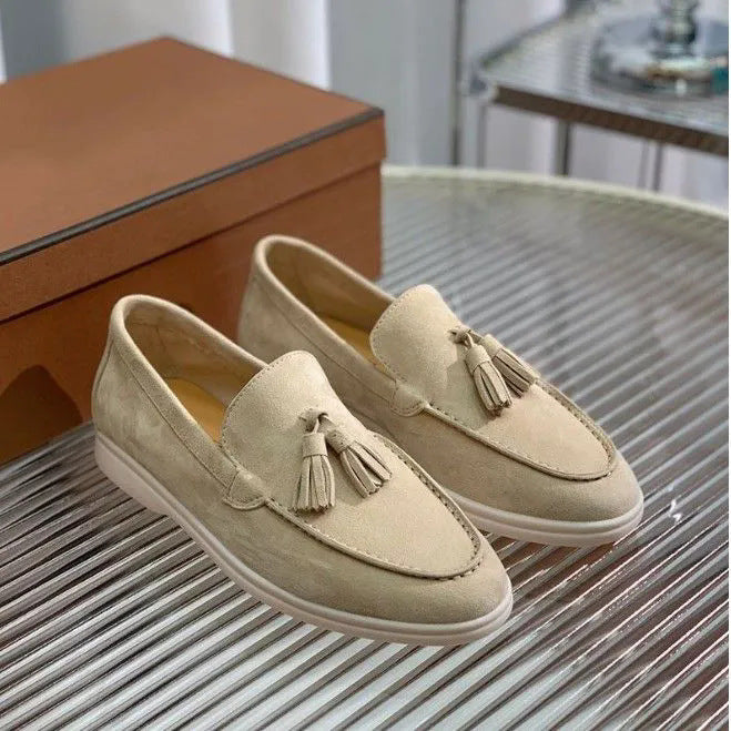 Mocassini beige in pelle scamosciata con nappine, suola bianca, su tavolo a righe, scatola marrone sullo sfondo. Scarpe eleganti uomo.