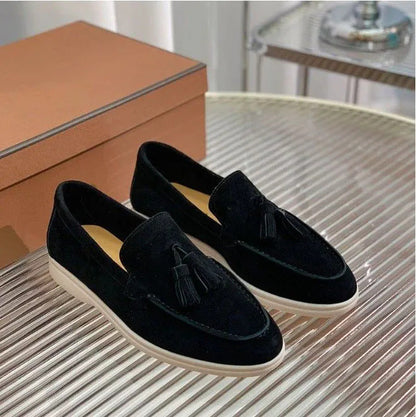 Mocassini neri in velluto con nappine su tavolo di vetro, scatola marrone sullo sfondo. Scarpe eleganti uomo, moda calzature di lusso.