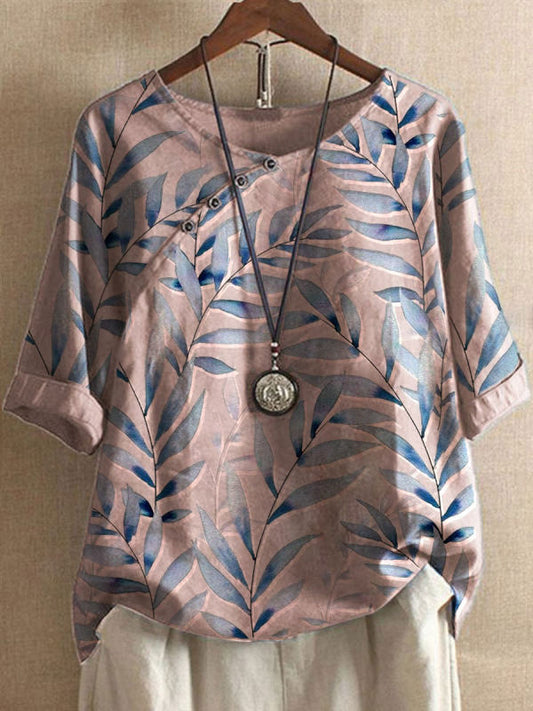 Maglietta casual da donna con stampa foglie blu su sfondo beige, maniche a tre quarti, abbinata a collana con pendente rotondo, appesa su gruccia.
