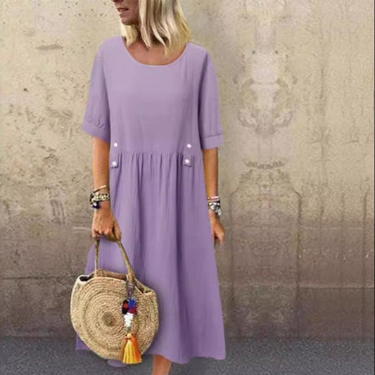 Donna con abito viola chiaro, maniche corte, tiene borsa di paglia rotonda con nappe colorate. Sfondo muro beige. Moda estiva casual.