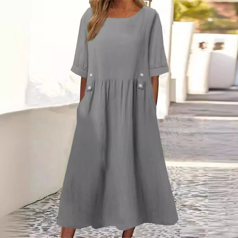 Abito lungo grigio da donna con maniche corte, tasche laterali e bottoni decorativi, ideale per look casual estivi. Sfondo urbano luminoso.