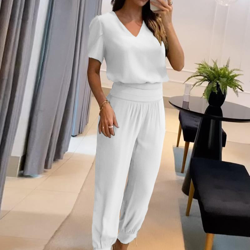 Donna in salotto moderno indossa elegante completo bianco, composto da maglia a maniche corte e pantaloni larghi. Tavolo nero con pianta decorativa.