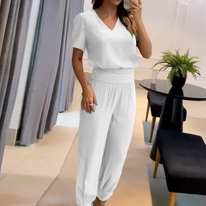 Donna in salotto moderno indossa elegante completo bianco, composto da maglia a maniche corte e pantaloni larghi. Tavolo nero con pianta decorativa.