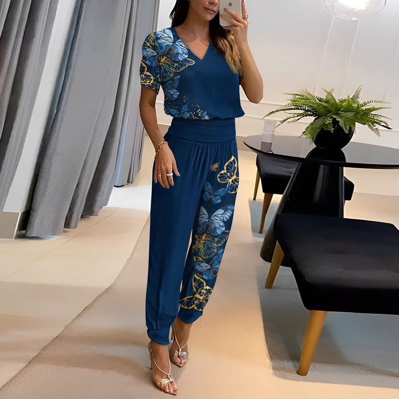 Donna in tuta blu con motivi floreali e farfalle, in piedi in un salotto moderno con tavolo nero e pianta decorativa. Moda elegante e casual.