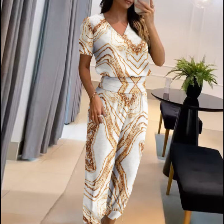 Donna in salotto moderno indossa tuta elegante bianca con motivi dorati. Tavolo nero e pianta decorativa sullo sfondo. Moda femminile chic.