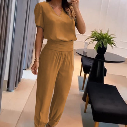 Donna in tuta elegante color senape, in piedi in un salotto moderno con tavolo nero e pianta decorativa. Moda femminile, abbigliamento casual chic.