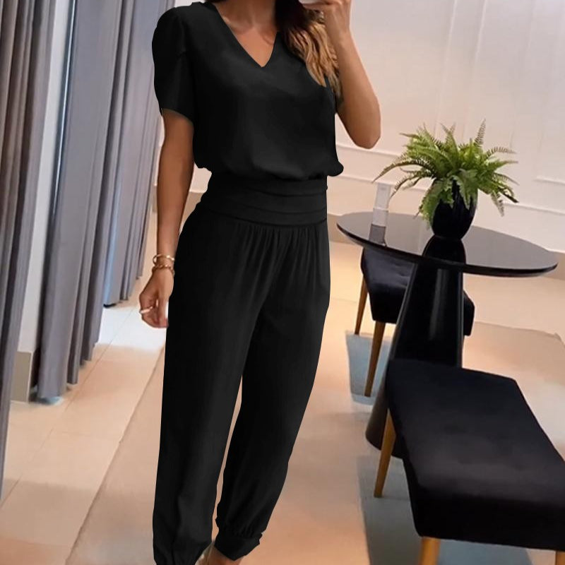 Donna in tuta nera elegante, con pantaloni larghi e maglia a maniche corte, in un moderno salotto con tavolo nero e pianta decorativa.