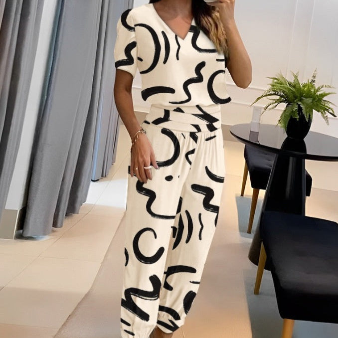 Donna in salotto indossa completo bianco con motivi astratti neri. Tavolo moderno con pianta, sedie nere. Design d'interni elegante e minimalista.