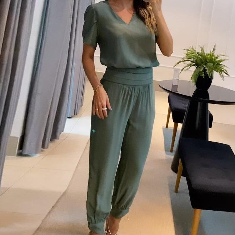 Donna in tuta verde elegante, con pantaloni a vita alta e maglia a maniche corte, in un ambiente moderno con tavolo nero e pianta decorativa.