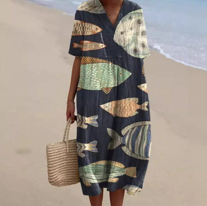 Donna in abito estivo con stampa pesci colorati, spiaggia sabbiosa sullo sfondo, borsa di paglia. Moda mare, abbigliamento estivo casual.