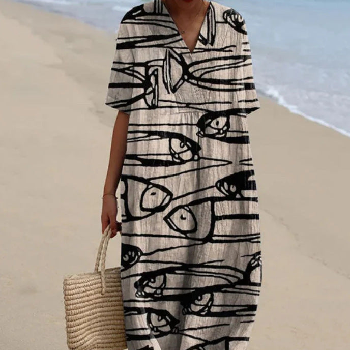 Donna in abito estivo con stampa astratta in bianco e nero, cammina su una spiaggia sabbiosa, tenendo una borsa di paglia intrecciata.
