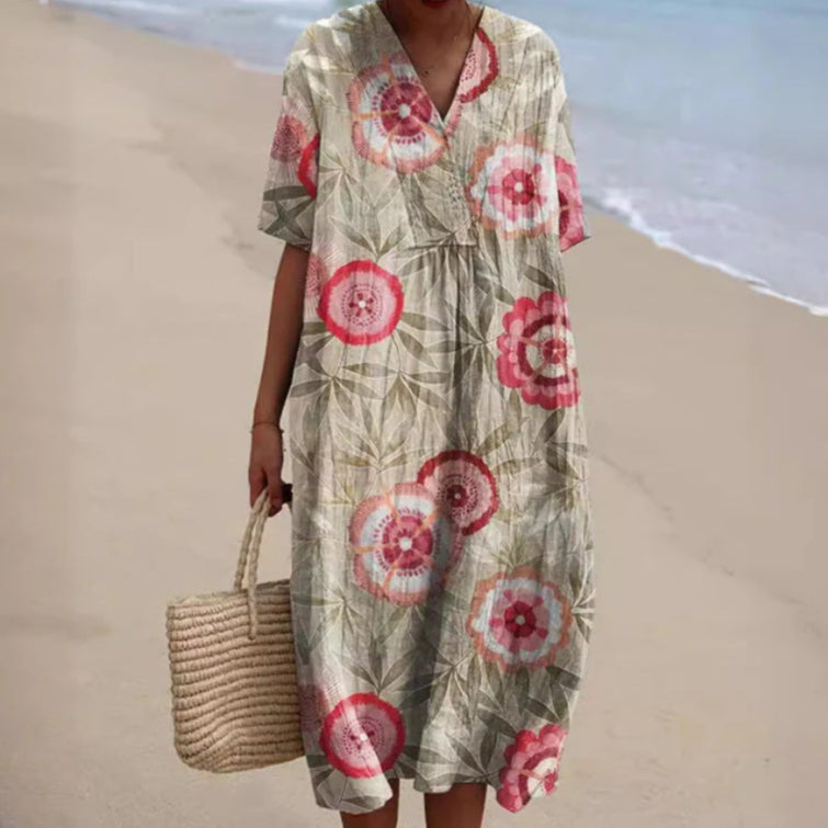 Abito estivo lungo con stampa floreale rossa e verde, indossato su spiaggia sabbiosa. Borsa di paglia intrecciata a mano. Moda mare donna.