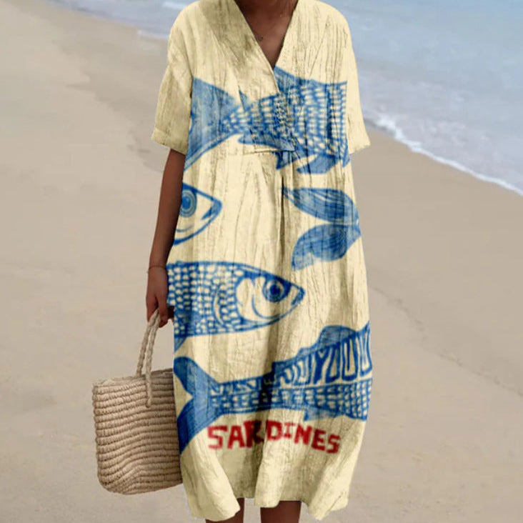 Donna in abito estivo beige con stampa blu di sardine, su spiaggia sabbiosa. Moda mare, abbigliamento casual, stile estivo.