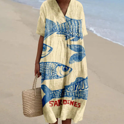 Donna in abito estivo beige con stampa blu di sardine, su spiaggia sabbiosa. Moda mare, abbigliamento casual, stile estivo.