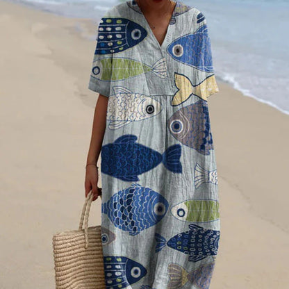 Donna in abito estivo con stampa di pesci colorati, cammina su una spiaggia sabbiosa. Moda mare, abbigliamento estivo, vacanza al mare.